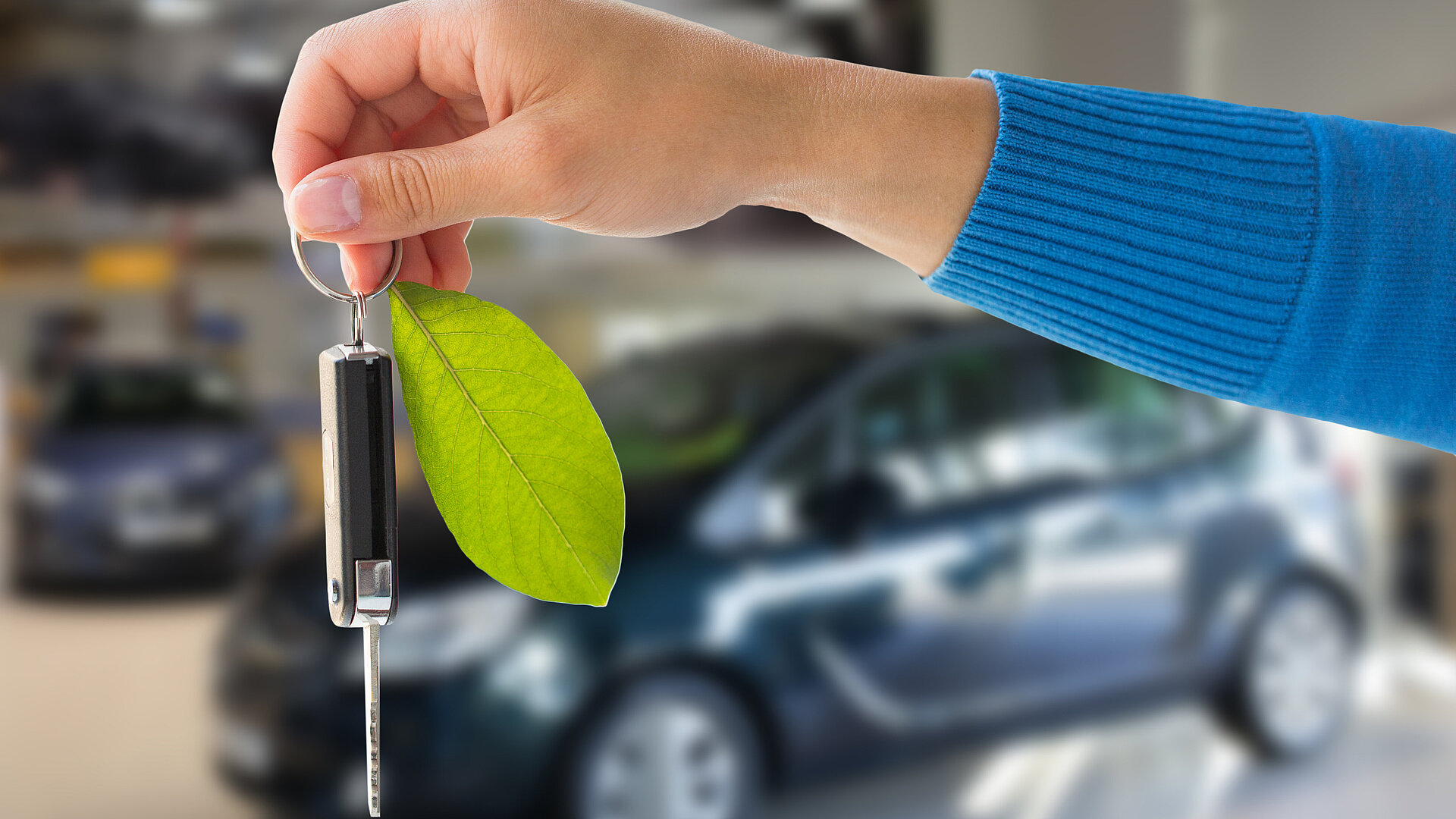 Hand hält Autoschlüssel mit grünem Blatt als Anhänger. Im Hintergund ist unscharf ein Auto zu sehen