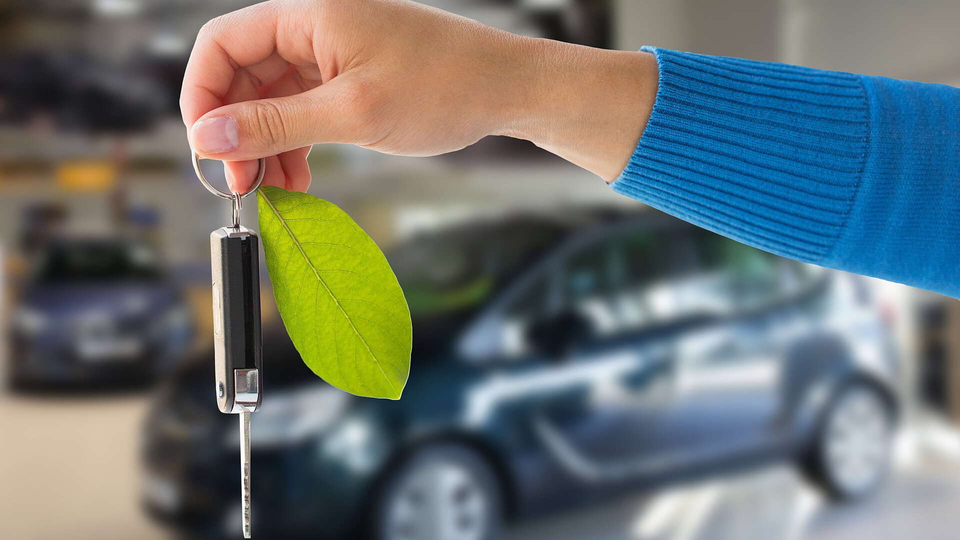 Hand hält Autoschlüssel mit grünem Blatt als Anhänger. Im Hintergund ist unscharf ein Auto zu sehen