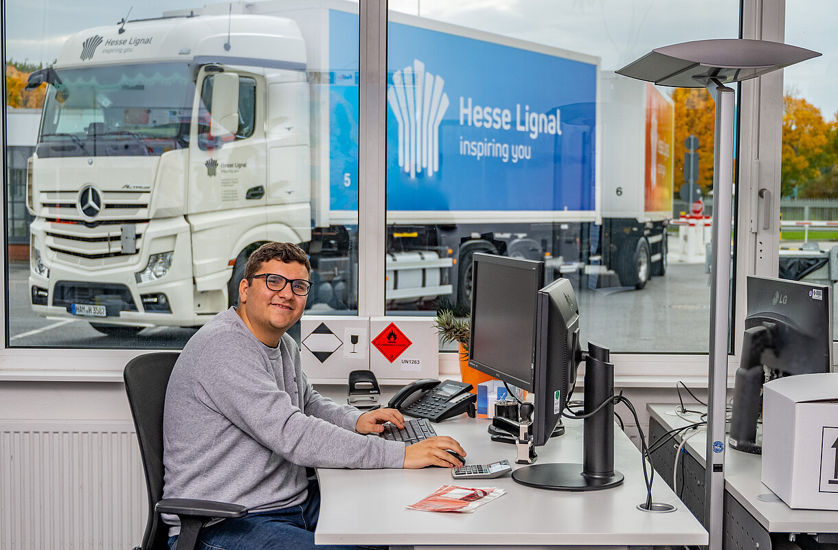 Kauffrau/-mann für Spedition und Logistikdienstleistungen am Schreibtisch und einem Hesse Lignal LKW im Hintergrund